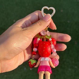 May include: A keychain featuring a small figure with a red strawberry-patterned head, white angel wings, and a pink skirt. The charm is decorated with various beads, including green and red strawberry shapes, and attaches to a gold heart-shaped ring.