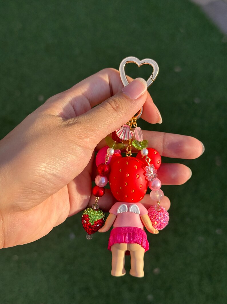 May include: A keychain featuring a small figure with a red strawberry-patterned head, white angel wings, and a pink skirt. The charm is decorated with various beads, including green and red strawberry shapes, and attaches to a gold heart-shaped ring.