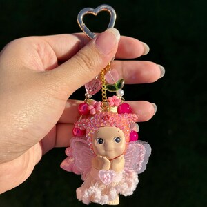 May include: A pink fairy keychain with a gold heart-shaped clasp. The fairy has a light skin tone, pink hair, and a pink dress. The keychain is adorned with pink beads, flowers, and a gold chain.