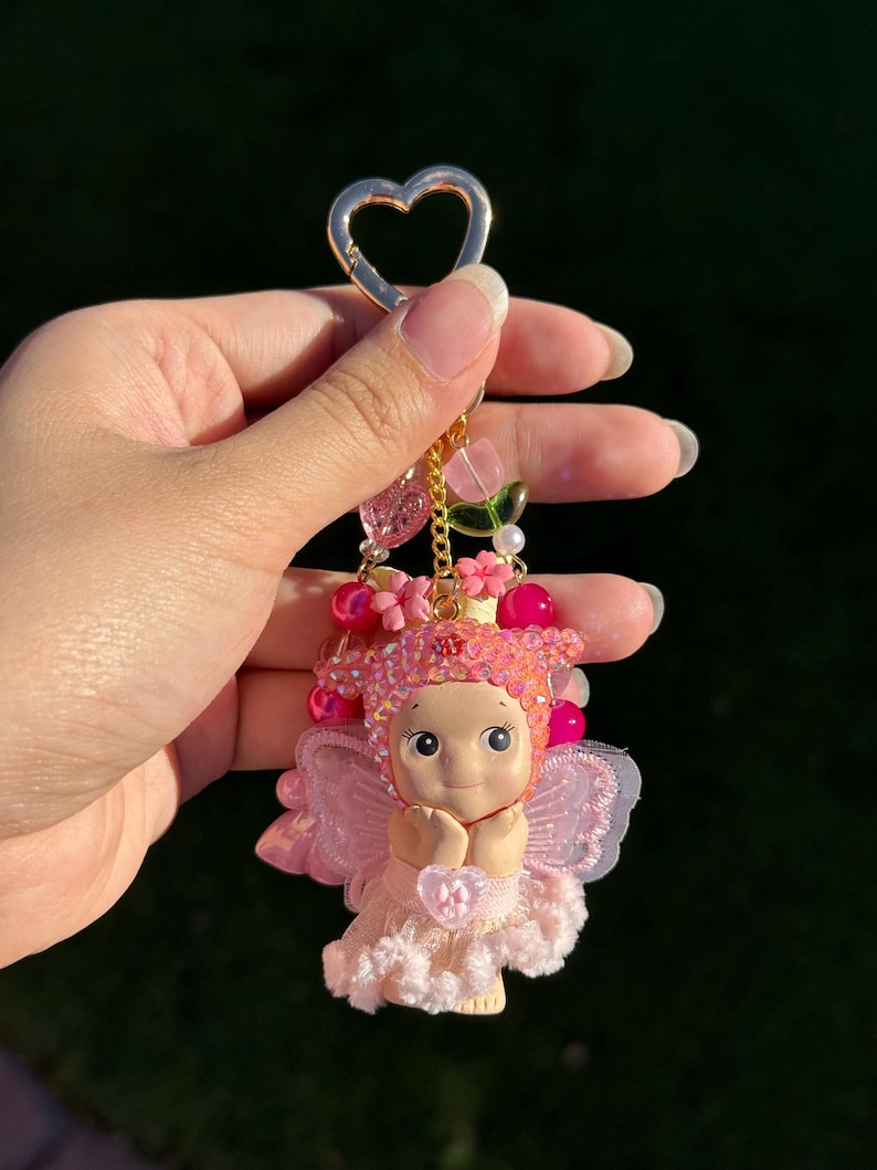 May include: A pink fairy keychain with a gold heart-shaped clasp. The fairy has a light skin tone, pink hair, and a pink dress. The keychain is adorned with pink beads, flowers, and a gold chain.
