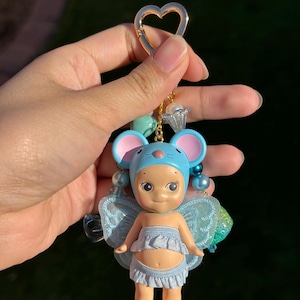 Puede incluir: Un llavero caprichoso con una peque&ntilde;a mu&ntilde;eca vestida de hada con un gorro de orejas de rat&oacute;n azul y alas. La mu&ntilde;eca tiene una falda con volantes azul claro y est&aacute; sujeta a un cierre dorado en forma de coraz&oacute;n con cuentas decorativas.