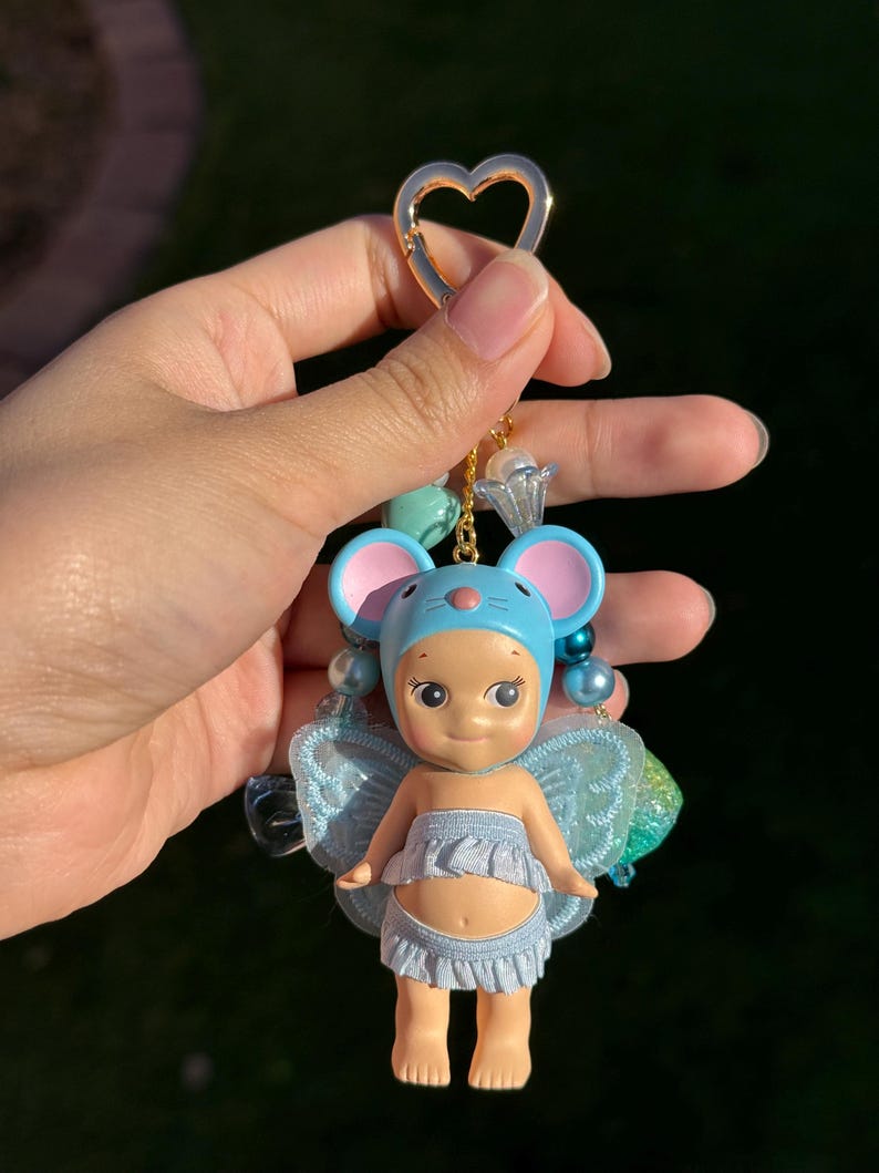 Puede incluir: Un llavero caprichoso con una peque&ntilde;a mu&ntilde;eca vestida de hada con un gorro de orejas de rat&oacute;n azul y alas. La mu&ntilde;eca tiene una falda con volantes azul claro y est&aacute; sujeta a un cierre dorado en forma de coraz&oacute;n con cuentas decorativas.