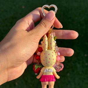 May include: A keychain featuring a pale yellow rabbit figure with pink spots, small white wings, and a bright pink skirt. The figure is attached to a gold-toned heart-shaped clasp and decorated with various beads, including red and pink spheres and a strawberry charm.