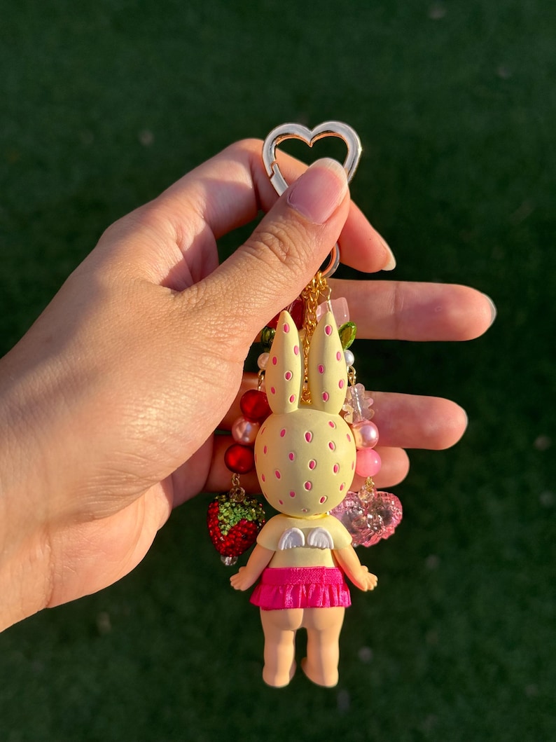 May include: A keychain featuring a pale yellow rabbit figure with pink spots, small white wings, and a bright pink skirt. The figure is attached to a gold-toned heart-shaped clasp and decorated with various beads, including red and pink spheres and a strawberry charm.
