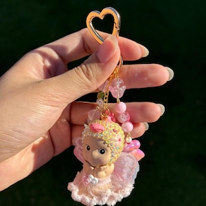 May include: A pink and gold keychain featuring a cherub figurine. The cherub has a pink and yellow jeweled headpiece, pink wings, and a pink dress. The keychain has a gold heart-shaped clasp and pink beaded accents.