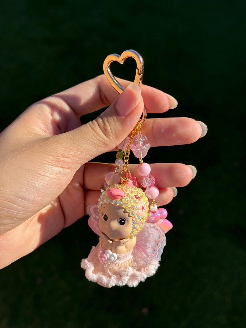 May include: A pink and gold keychain featuring a cherub figurine. The cherub has a pink and yellow jeweled headpiece, pink wings, and a pink dress. The keychain has a gold heart-shaped clasp and pink beaded accents.