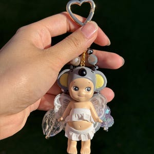 Puede incluir: Un llavero con una peque&ntilde;a mu&ntilde;eca vestida de koala con capucha gris y orejas amarillas. La mu&ntilde;eca lleva un top y una parte inferior con volantes blancos, con alas transparentes e iridiscentes. El llavero tiene un cierre dorado en forma de coraz&oacute;n y detalles de cuentas.