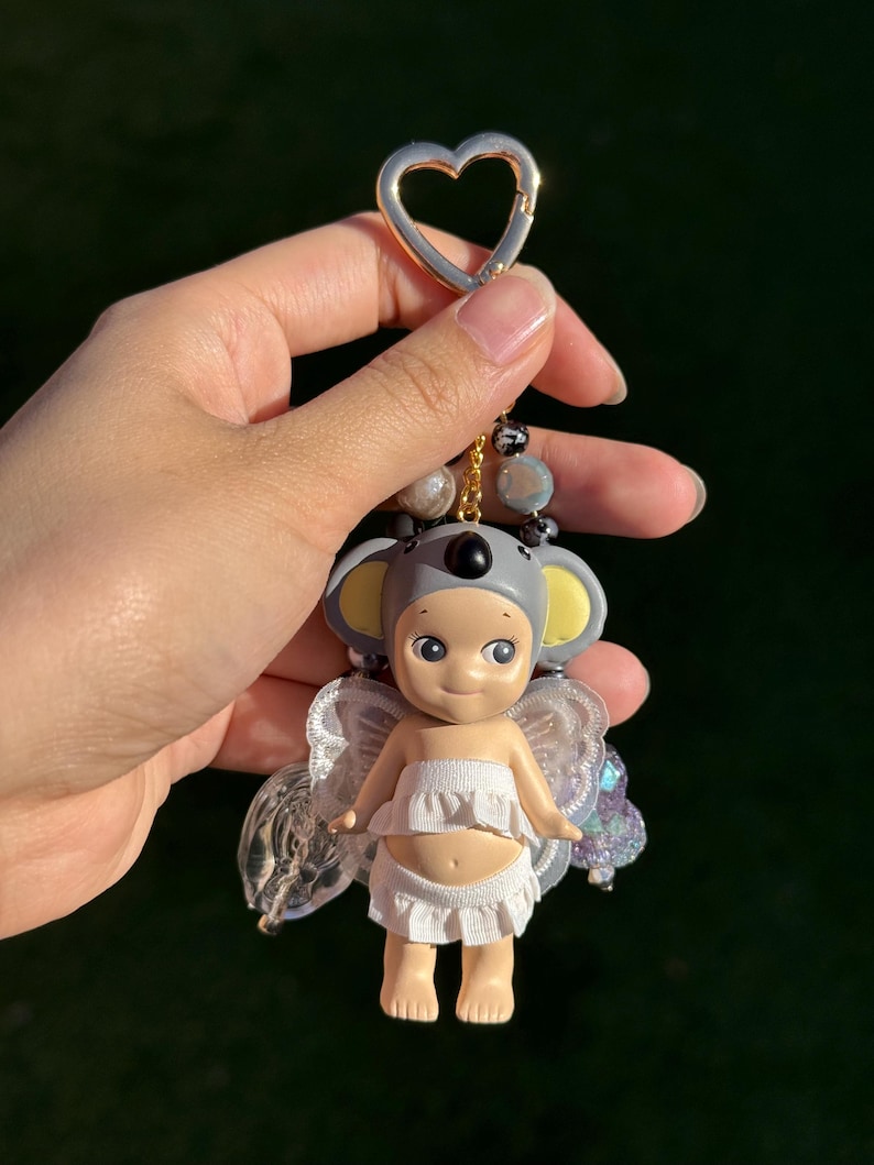 Puede incluir: Un llavero con una peque&ntilde;a mu&ntilde;eca vestida de koala con capucha gris y orejas amarillas. La mu&ntilde;eca lleva un top y una parte inferior con volantes blancos, con alas transparentes e iridiscentes. El llavero tiene un cierre dorado en forma de coraz&oacute;n y detalles de cuentas.