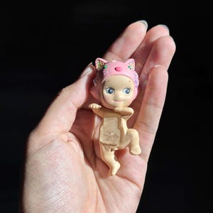May include: A small, light-skinned figurine wearing a pink cat-themed headpiece with iridescent accents. The figure has a neutral expression and a translucent, rectangular chest plate featuring the embossed text "DREAMS CHINA".