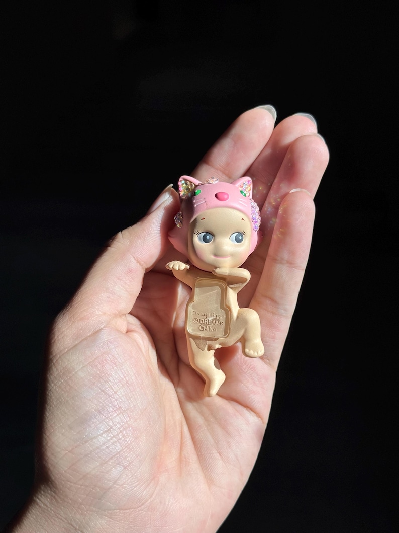 May include: A small, light-skinned figurine wearing a pink cat-themed headpiece with iridescent accents. The figure has a neutral expression and a translucent, rectangular chest plate featuring the embossed text "DREAMS CHINA".