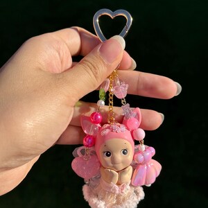 May include: A pink keychain featuring a small doll with a heart-shaped clasp. The doll has a pink bonnet, dress, and wings, with beaded accents in pink and clear. The keychain is held by a hand against a dark background.