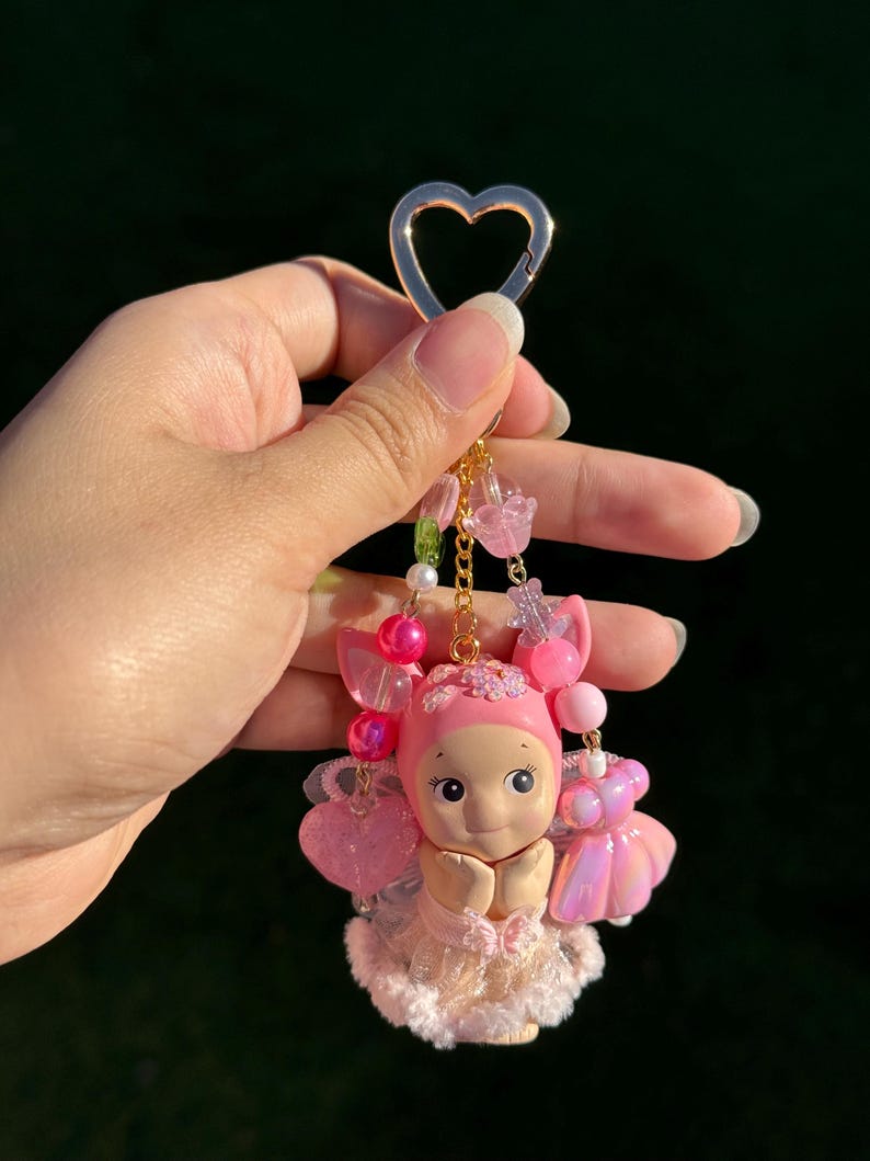 May include: A pink keychain featuring a small doll with a heart-shaped clasp. The doll has a pink bonnet, dress, and wings, with beaded accents in pink and clear. The keychain is held by a hand against a dark background.