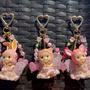 May include: Three keychains with gold heart-shaped clasps. Each keychain features a small doll with pink fairy wings and a matching outfit. One doll wears a bunny ear bonnet. The keychains are adorned with pink and pearl beads.