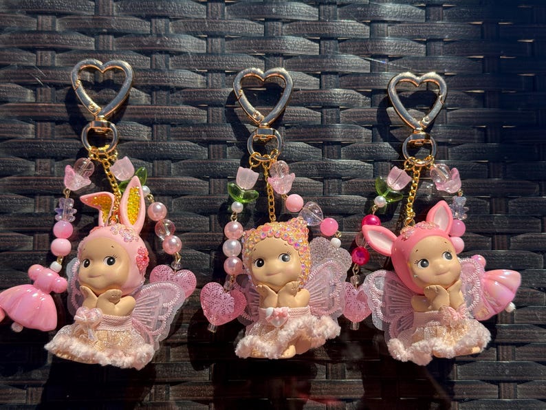 May include: Three keychains with gold heart-shaped clasps. Each keychain features a small doll with pink fairy wings and a matching outfit. One doll wears a bunny ear bonnet. The keychains are adorned with pink and pearl beads.