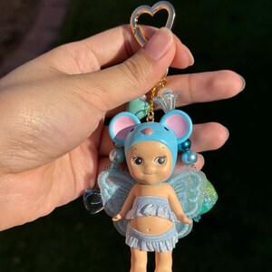 Puede incluir: Llavero de mu&ntilde;eca con tem&aacute;tica de rat&oacute;n azul y rosa con un cierre dorado en forma de coraz&oacute;n. La mu&ntilde;eca tiene un gorro azul, una falda con volantes y peque&ntilde;as alas azules. El llavero est&aacute; adornado con cuentas azules y transparentes.