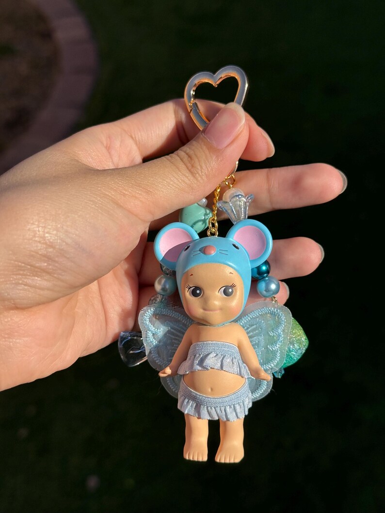 Puede incluir: Llavero de mu&ntilde;eca con tem&aacute;tica de rat&oacute;n azul y rosa con un cierre dorado en forma de coraz&oacute;n. La mu&ntilde;eca tiene un gorro azul, una falda con volantes y peque&ntilde;as alas azules. El llavero est&aacute; adornado con cuentas azules y transparentes.