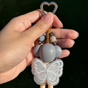 Puede incluir: Un llavero de figura gris con un dise&ntilde;o de encaje en forma de mariposa. El llavero tiene un cierre dorado en forma de coraz&oacute;n y una cadena de cuentas con cuentas grises, azules y marrones. La figura tiene una base marr&oacute;n claro y una cabeza gris.