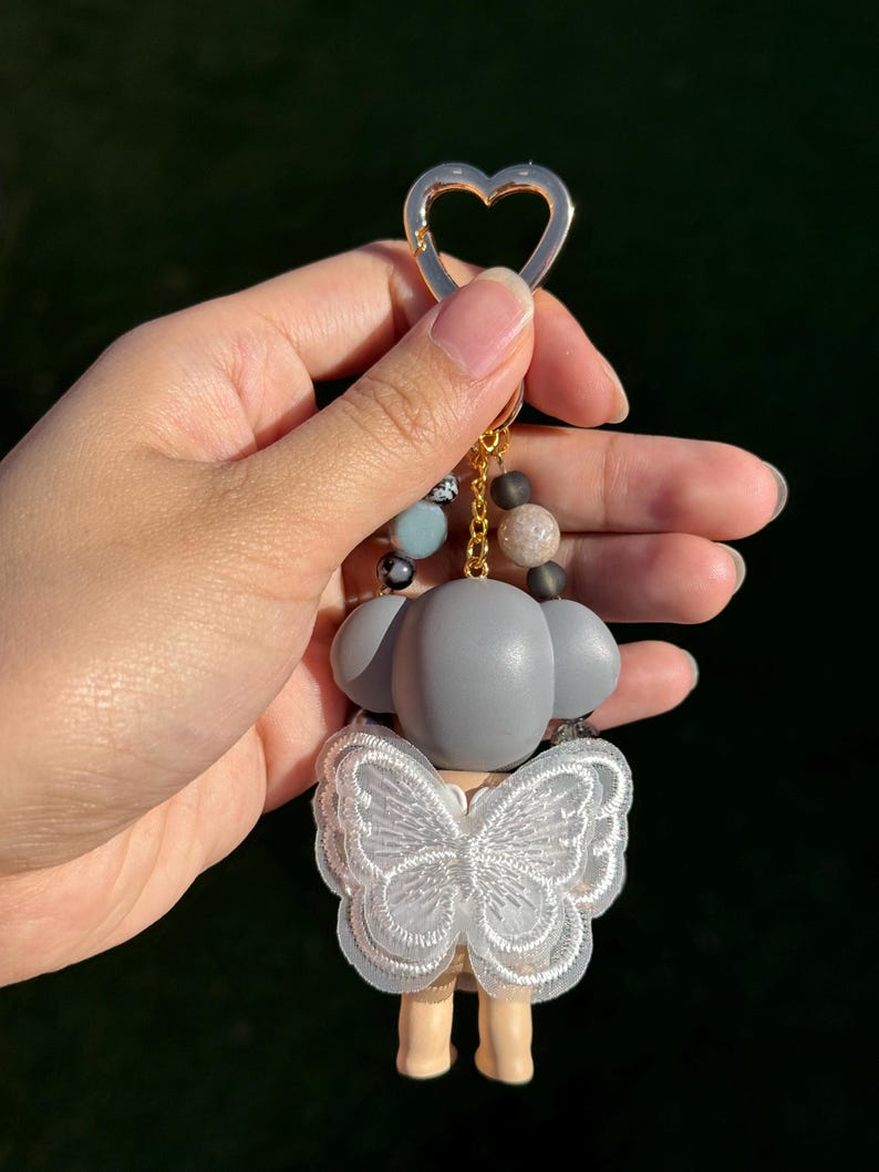 Puede incluir: Un llavero de figura gris con un dise&ntilde;o de encaje en forma de mariposa. El llavero tiene un cierre dorado en forma de coraz&oacute;n y una cadena de cuentas con cuentas grises, azules y marrones. La figura tiene una base marr&oacute;n claro y una cabeza gris.