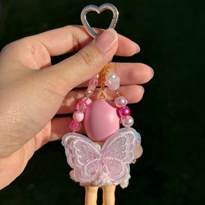 Puede incluir: Un llavero rosa y blanco con un cierre dorado en forma de coraz&oacute;n. El llavero presenta una mu&ntilde;eca rosa con un dise&ntilde;o de alas de mariposa y peque&ntilde;as piernas amarillas. El llavero es sostenido por una mano sobre un fondo verde borroso.