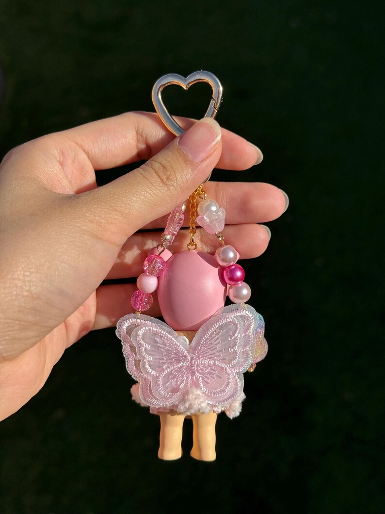 Puede incluir: Un llavero rosa y blanco con un cierre dorado en forma de coraz&oacute;n. El llavero presenta una mu&ntilde;eca rosa con un dise&ntilde;o de alas de mariposa y peque&ntilde;as piernas amarillas. El llavero es sostenido por una mano sobre un fondo verde borroso.