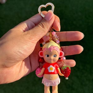 May include: A keychain featuring a small figurine with blonde hair, a pink strawberry-patterned cap, a red shirt with a white flower, and a light pink skirt. The charm is accented with pink and red beads and a red strawberry-shaped pendant.