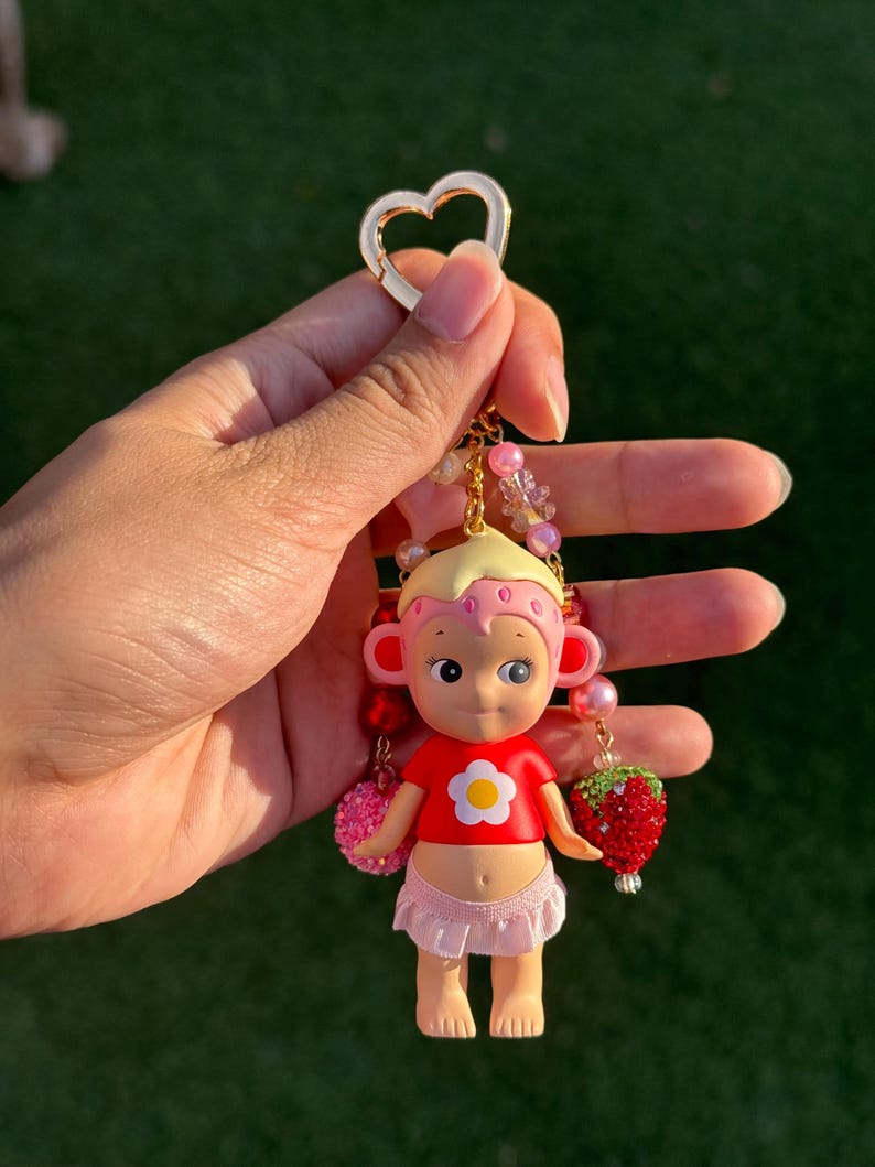 May include: A keychain featuring a small figurine with blonde hair, a pink strawberry-patterned cap, a red shirt with a white flower, and a light pink skirt. The charm is accented with pink and red beads and a red strawberry-shaped pendant.