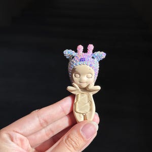 May include: A small figurine with a smooth beige body and a head covered in iridescent, multi-colored sequins. The figure has a rounded face, closed eyes, and hands positioned near the cheeks. A clear, rectangular plastic insert is on the torso.