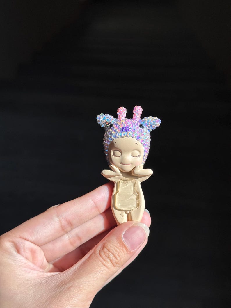 May include: A small figurine with a smooth beige body and a head covered in iridescent, multi-colored sequins. The figure has a rounded face, closed eyes, and hands positioned near the cheeks. A clear, rectangular plastic insert is on the torso.