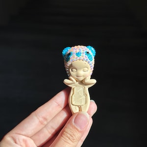 May include: A small, beige figurine of a character with closed eyes and hands near the face. The character wears a headpiece covered in iridescent, light-reflecting beads with bright blue accents on the ears and forehead area.