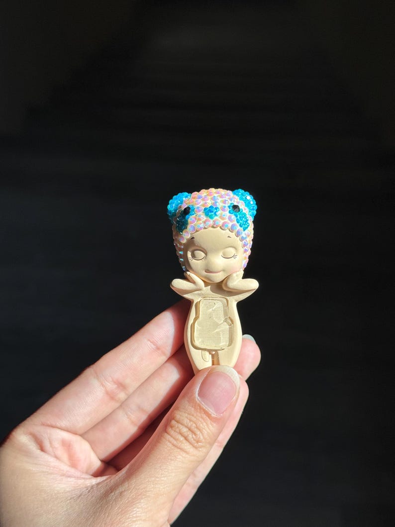 May include: A small, beige figurine of a character with closed eyes and hands near the face. The character wears a headpiece covered in iridescent, light-reflecting beads with bright blue accents on the ears and forehead area.