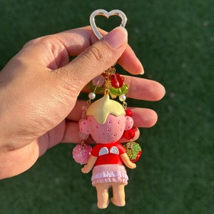 May include: A keychain featuring a small doll with a pink strawberry-textured head, a yellow cap, and a red shirt with white wings. The doll wears a light pink ruffled skirt and is surrounded by dangling beads, including red and green fruit charms.