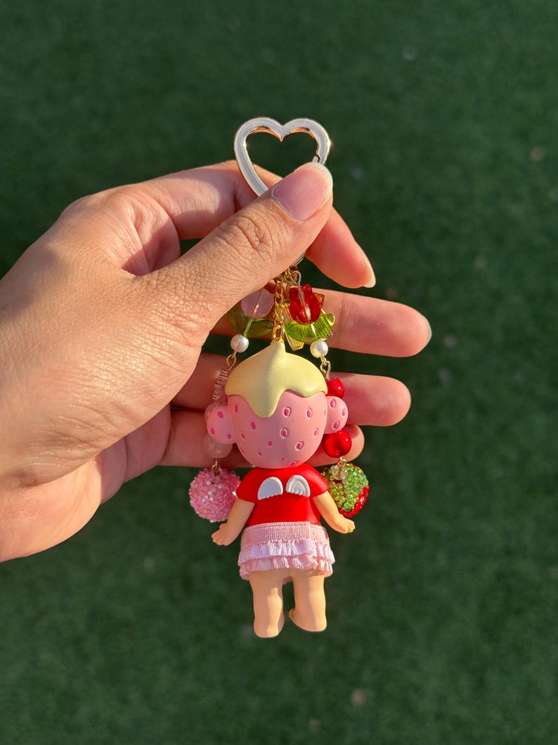 May include: A keychain featuring a small doll with a pink strawberry-textured head, a yellow cap, and a red shirt with white wings. The doll wears a light pink ruffled skirt and is surrounded by dangling beads, including red and green fruit charms.