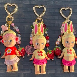 May include: Five keychains featuring small figurines with various headgear, including rabbit ears and animal motifs. Each figure wears a colorful top and skirt, accented with beaded strands, strawberry charms, and gold-toned heart-shaped clasps.