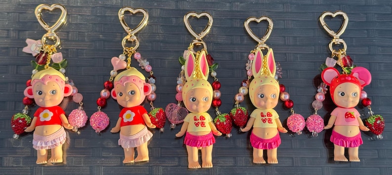 May include: Five keychains featuring small figurines with various headgear, including rabbit ears and animal motifs. Each figure wears a colorful top and skirt, accented with beaded strands, strawberry charms, and gold-toned heart-shaped clasps.
