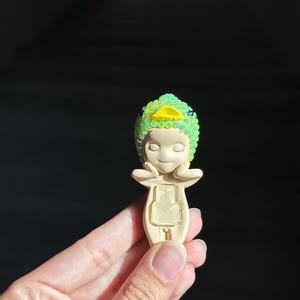 May include: A small, beige figurine featuring a character wearing a bright green, iridescent, sequined duck-shaped hood with a yellow beak. The figure has closed eyes, hands resting near its face, and a rectangular cutout on its torso.
