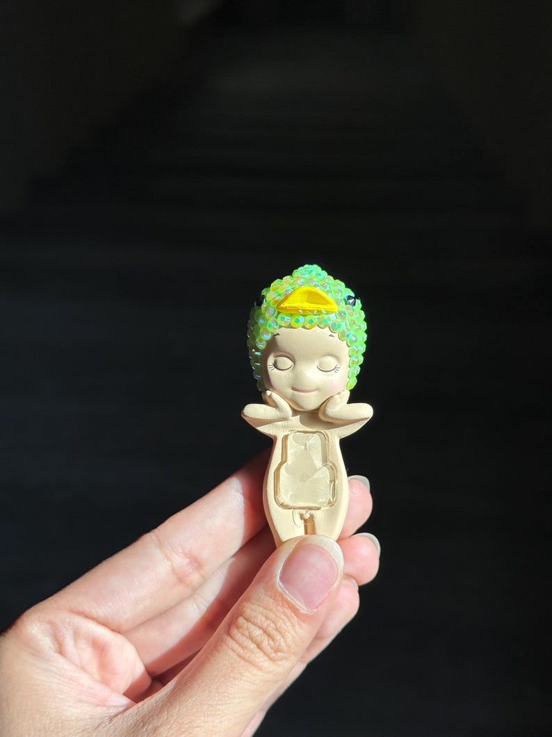 May include: A small, beige figurine featuring a character wearing a bright green, iridescent, sequined duck-shaped hood with a yellow beak. The figure has closed eyes, hands resting near its face, and a rectangular cutout on its torso.