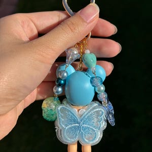Puede incluir: Un llavero hecho a mano con una peque&ntilde;a mu&ntilde;eca con cabeza azul claro y un vestido con forma de mariposa. El llavero incluye un cierre dorado en forma de coraz&oacute;n y est&aacute; adornado con varias cuentas azules y transparentes. Las piernas de la mu&ntilde;eca son de color marr&oacute;n claro.