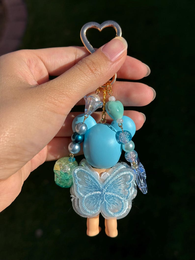 Puede incluir: Un llavero hecho a mano con una peque&ntilde;a mu&ntilde;eca con cabeza azul claro y un vestido con forma de mariposa. El llavero incluye un cierre dorado en forma de coraz&oacute;n y est&aacute; adornado con varias cuentas azules y transparentes. Las piernas de la mu&ntilde;eca son de color marr&oacute;n claro.