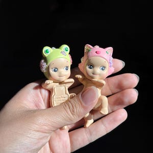 May include: Two small collectible figurines with light skin tones. One wears a light green frog-themed headpiece, and the other wears a pink cat-themed headpiece. Both feature iridescent, sparkling accents on the sides of their headgear.