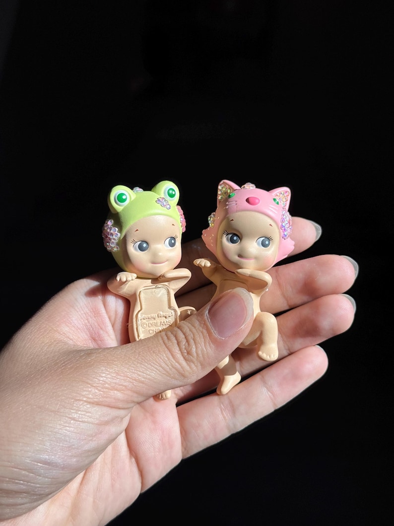 May include: Two small collectible figurines with light skin tones. One wears a light green frog-themed headpiece, and the other wears a pink cat-themed headpiece. Both feature iridescent, sparkling accents on the sides of their headgear.
