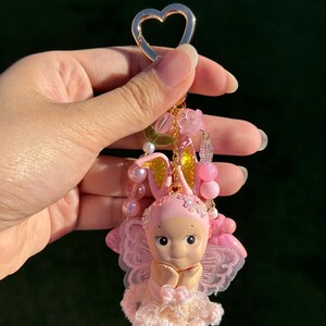 May include: A pink fairy keychain with a gold heart-shaped clasp. The fairy figurine has a pink bonnet with gold ears, a lace dress, and delicate wings. The keychain is adorned with pink beads and flower accents.