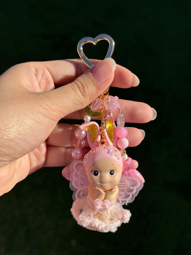 May include: A pink fairy keychain with a gold heart-shaped clasp. The fairy figurine has a pink bonnet with gold ears, a lace dress, and delicate wings. The keychain is adorned with pink beads and flower accents.
