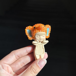 May include: A small figurine featuring a smooth, light-colored body with hands held near the face. The head is adorned with a textured, orange, crystal-covered elephant-shaped hood, featuring large ears with bright blue accents on the inner sides.