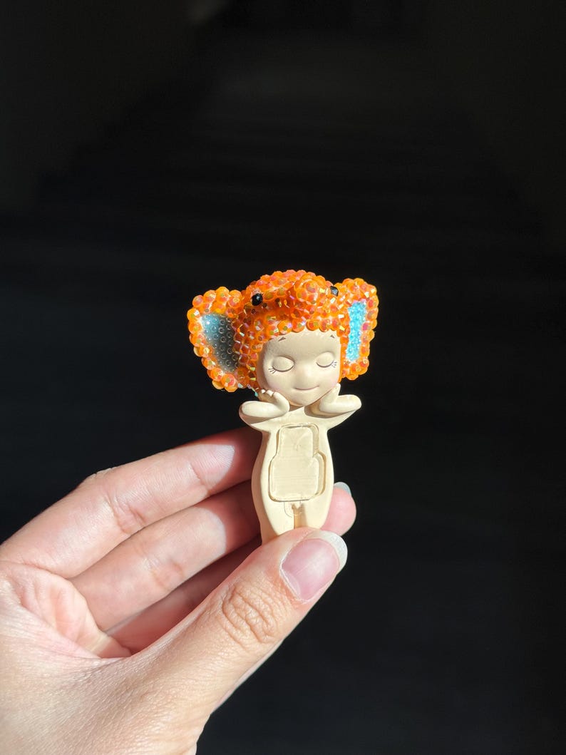 May include: A small figurine featuring a smooth, light-colored body with hands held near the face. The head is adorned with a textured, orange, crystal-covered elephant-shaped hood, featuring large ears with bright blue accents on the inner sides.