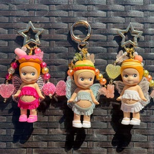 Puede incluir: Cinco llaveros con pequeñas figuras de muñecas aladas, cada una con un atuendo y accesorios únicos. Las figuras tienen cabello naranja y están unidas a dijes en forma de estrella. Los atuendos incluyen vestidos, faldas y sombreros, con detalles de cuentas de colores.