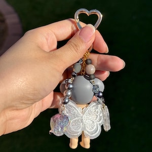 Puede incluir: Un llavero decorativo con una peque&ntilde;a figura. La figura tiene una cabeza gris y un vestido de mariposa de encaje blanco. El llavero tiene un cierre dorado en forma de coraz&oacute;n y est&aacute; adornado con cuentas grises, marrones y cristales transparentes.