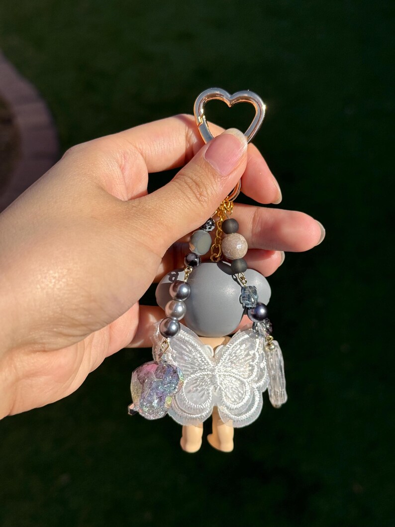 Puede incluir: Un llavero decorativo con una peque&ntilde;a figura. La figura tiene una cabeza gris y un vestido de mariposa de encaje blanco. El llavero tiene un cierre dorado en forma de coraz&oacute;n y est&aacute; adornado con cuentas grises, marrones y cristales transparentes.