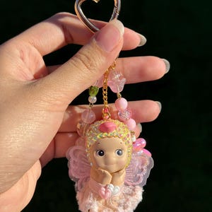 May include: A pink and gold keychain featuring a cherubic doll with a heart-shaped charm. The doll has a pink dress, wings, and a sequined headpiece. The keychain includes pink and green beads and a gold heart-shaped clasp.