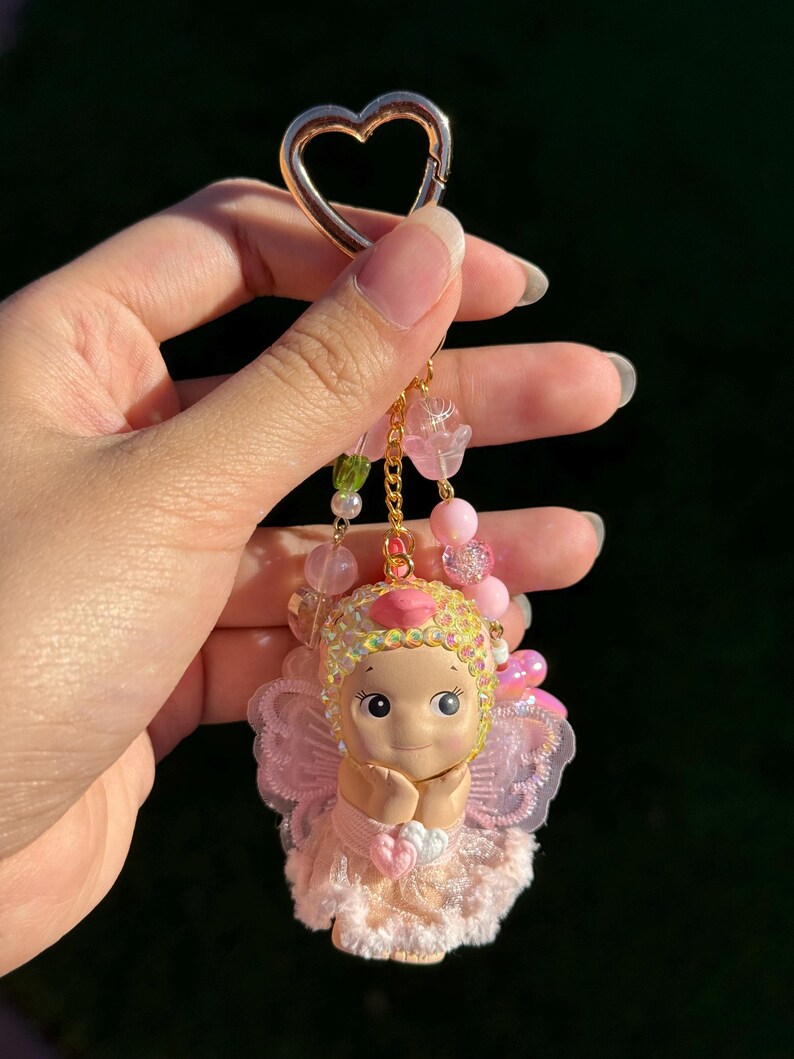 May include: A pink and gold keychain featuring a cherubic doll with a heart-shaped charm. The doll has a pink dress, wings, and a sequined headpiece. The keychain includes pink and green beads and a gold heart-shaped clasp.
