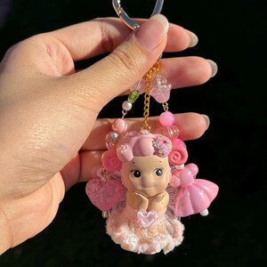 May include: A pink and gold keychain featuring a small doll with a heart-shaped charm. The doll is wearing a pink dress and has pink hair. The keychain also includes pink beads and a gold heart-shaped clasp.